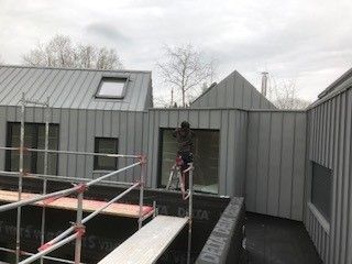 Eine Person arbeitet auf einer Leiter an der Außenfassade eines Gebäudes, das mit grauen Metallpaneelen verkleidet ist.