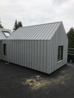 Graue, metallverkleidete, schuppenartige Konstruktion mit Satteldach und zwei kleinen Fenstern auf einem Flachdach.