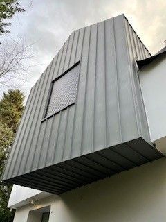 Architektonisches Element mit grauer Metallverkleidung, Fenster und überhängendem Teil, das an ein weißes Gebäude angebaut ist.