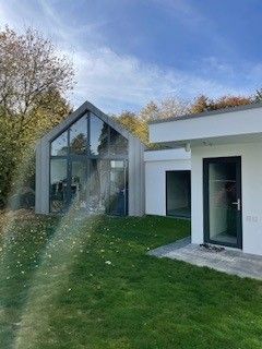 Modernes Haus mit giebelständiger Glasfassade, das mit einem weißen Anbau mit schwarzen Türen verbunden ist, auf einem grünen Rasen.