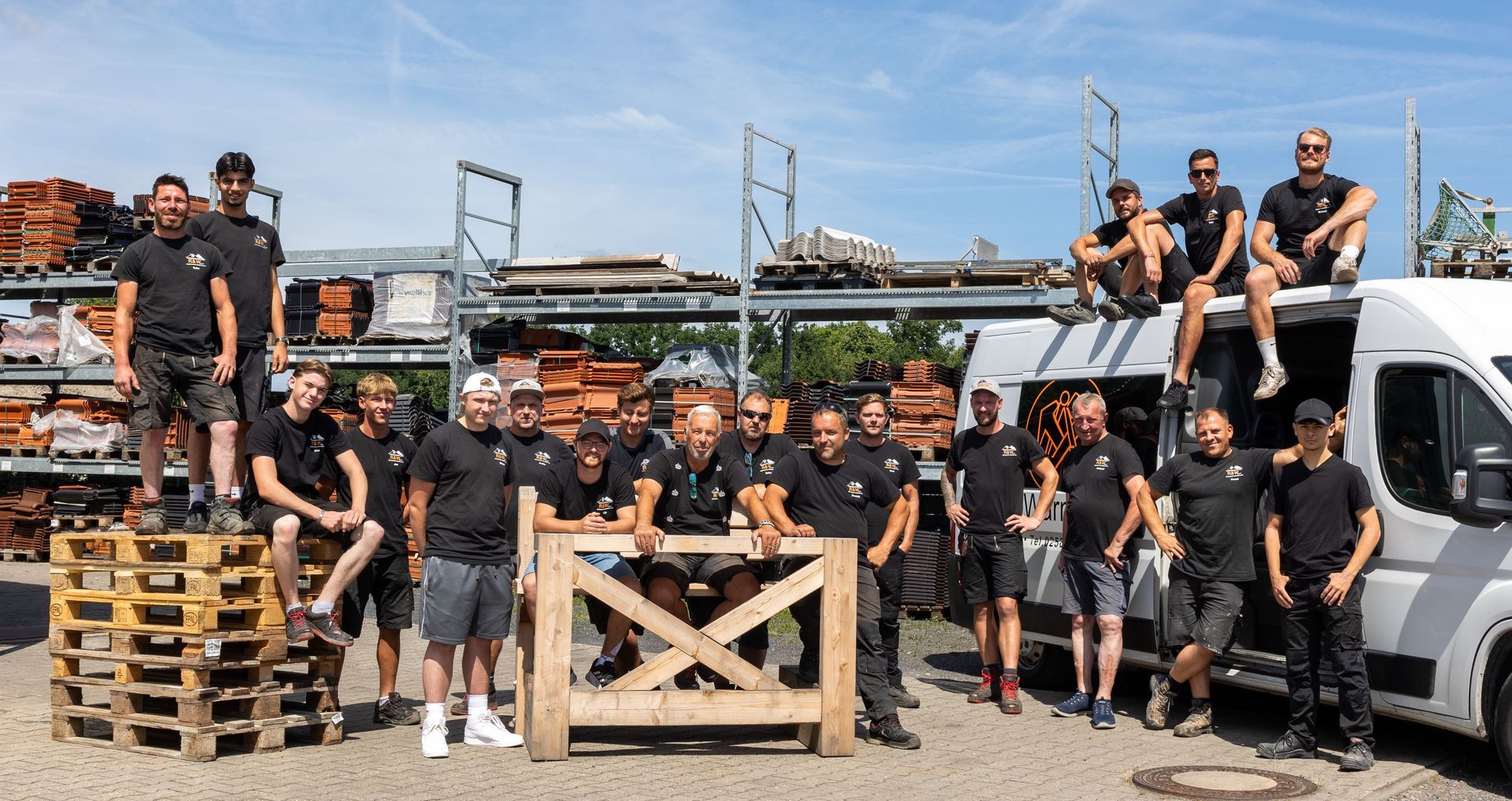 Eine Gruppe von Bauarbeitern posiert draußen mit Baugeräten; einige stehen auf Paletten und einem Lieferwagen.