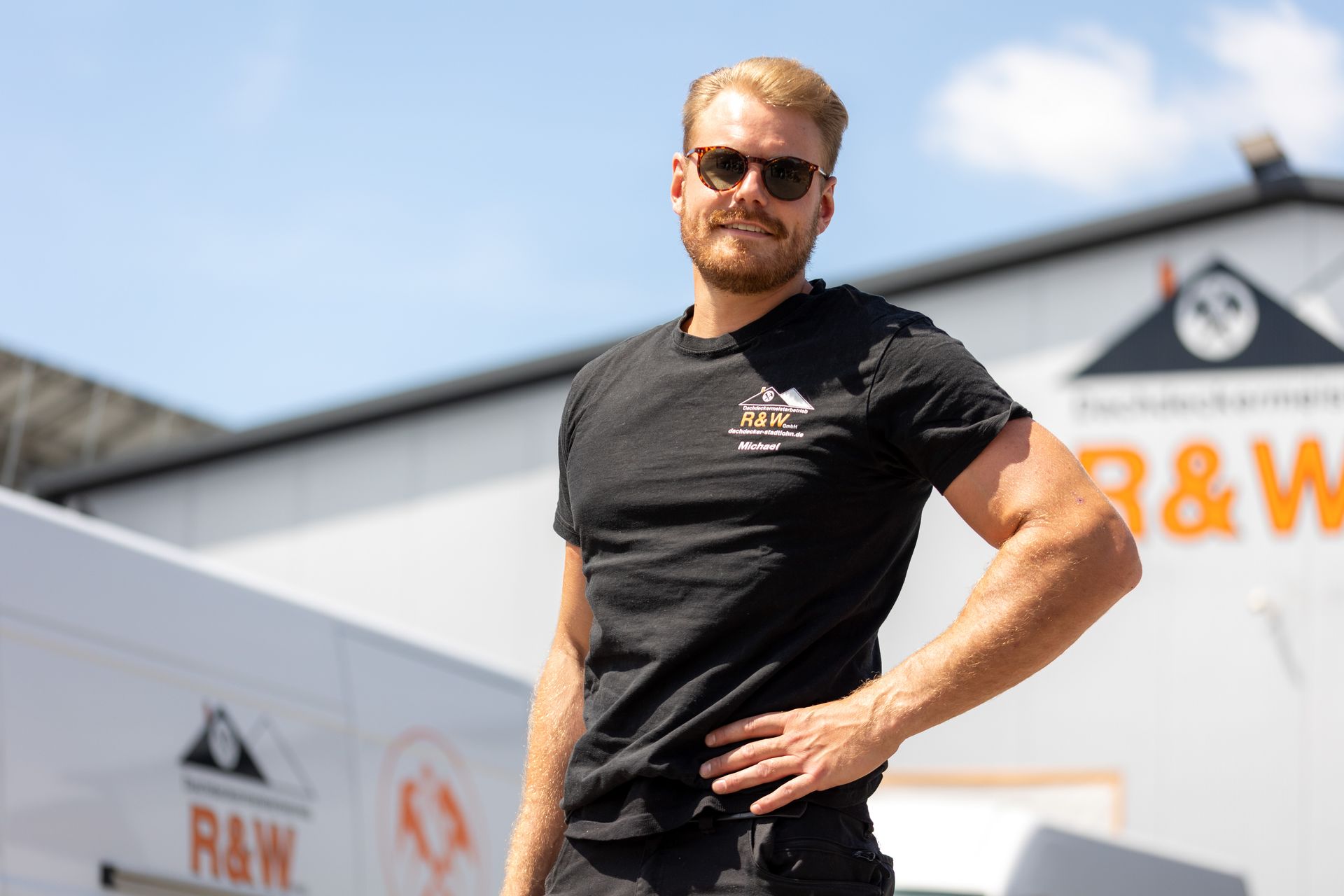 Ein Mann mit Sonnenbrille und schwarzem T-Shirt steht vor einem Gebäude mit dem Logo „R&W“.