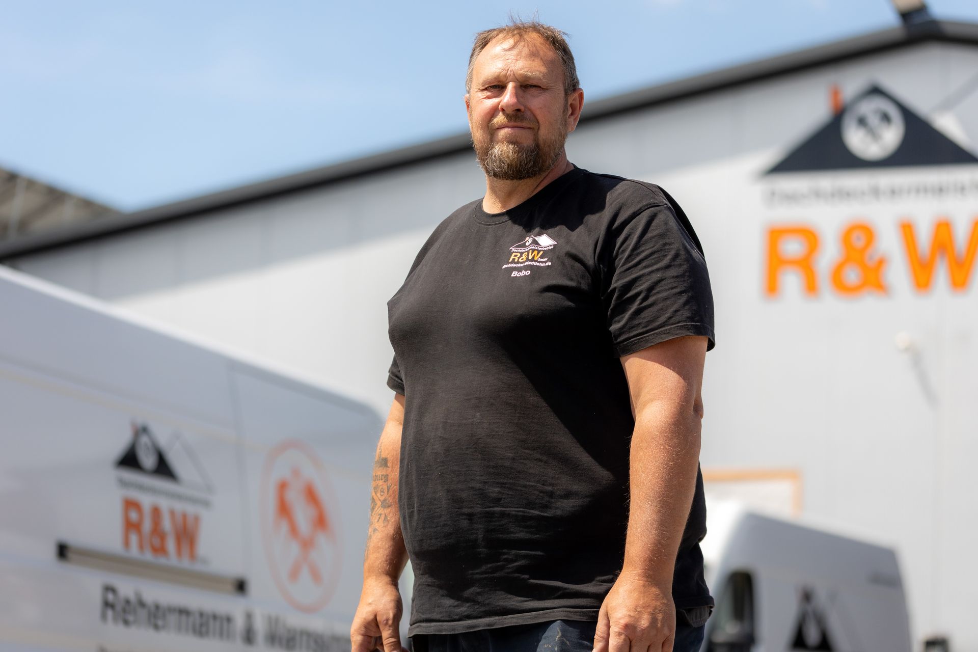 Ein Mann in schwarzem Hemd steht vor einem Gebäude und einem Lieferwagen mit dem „R&W“-Logo. Sonniger Tag.