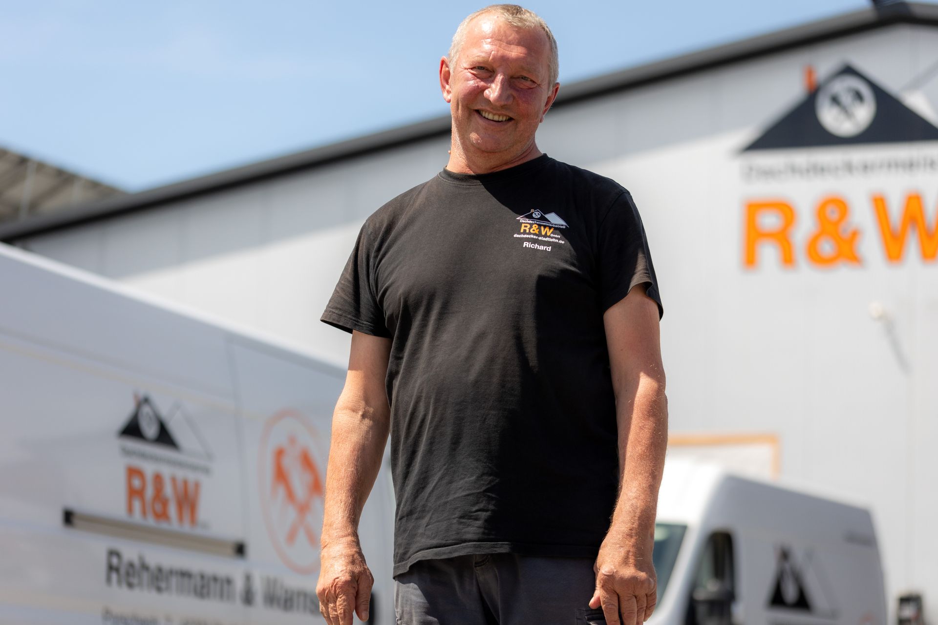 Ein Mann in schwarzem Hemd lächelt vor weißen Lieferwagen und einem Gebäude mit dem „R&W“-Logo.