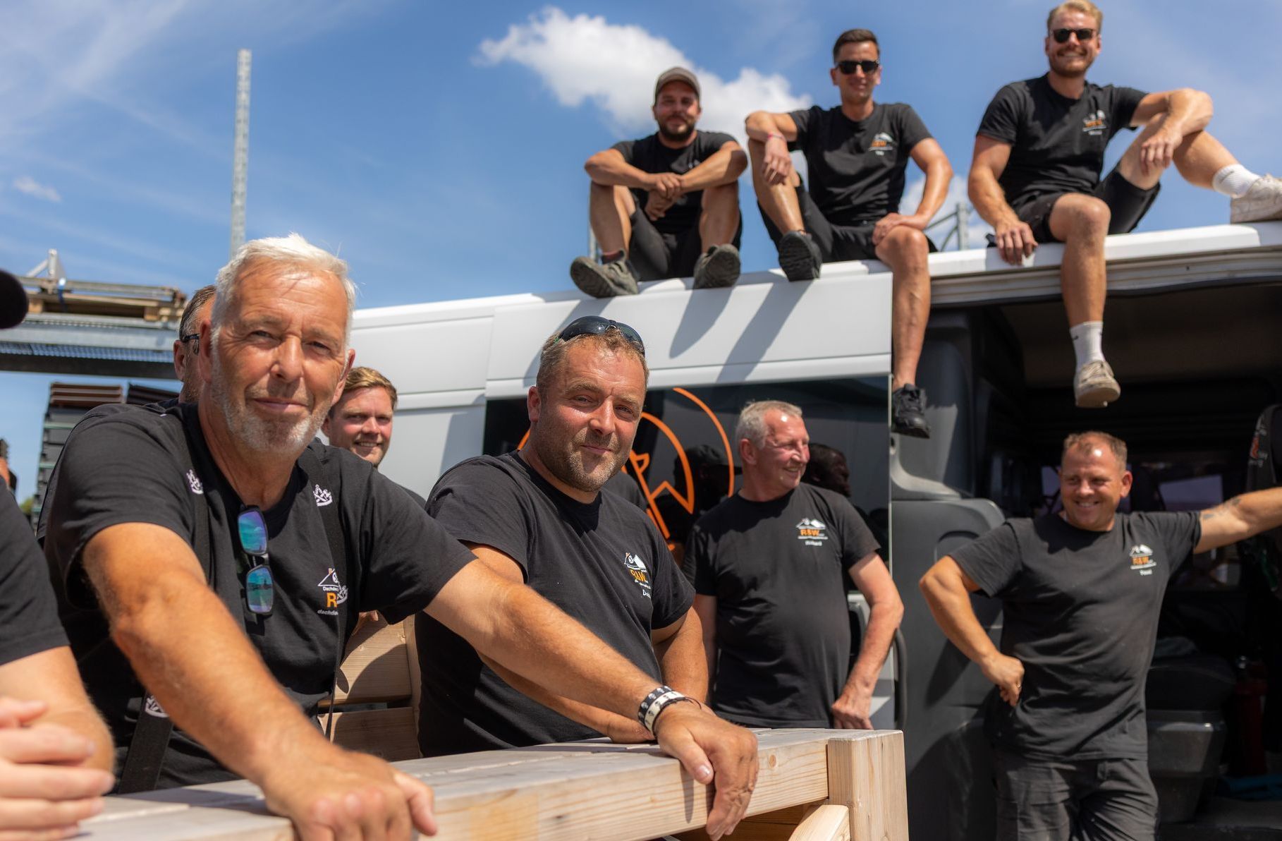 Eine Gruppe Männer in schwarzen Hemden posiert an einem sonnigen Tag neben einem Lieferwagen. Einige sitzen auf dem Dach.