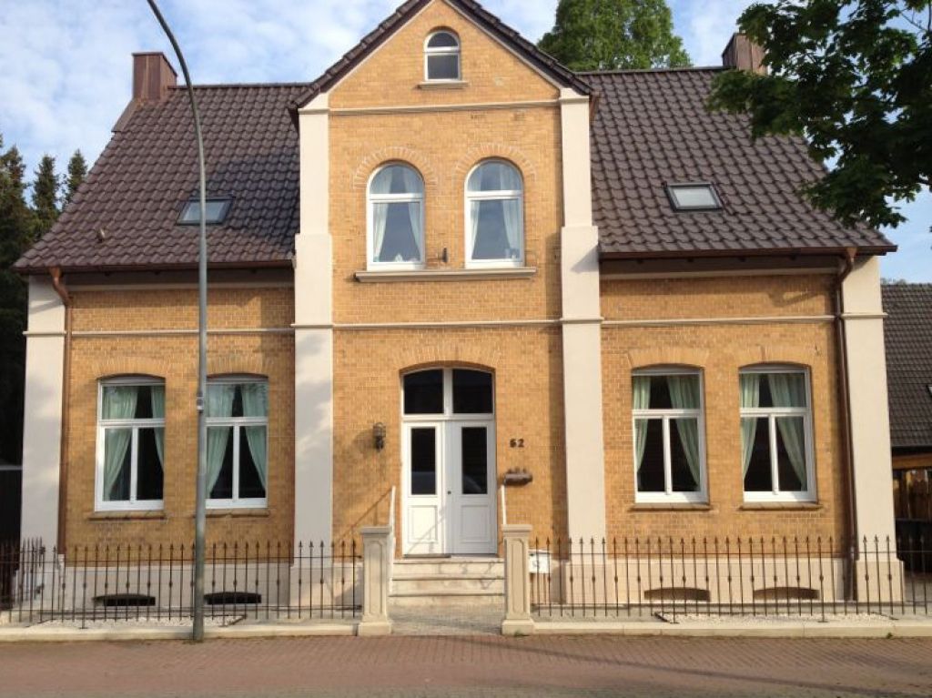 Beiges Haus mit braunem Dach und weißen Zierelementen. Weiße Tür und Fenster, schwarzer Zaun.