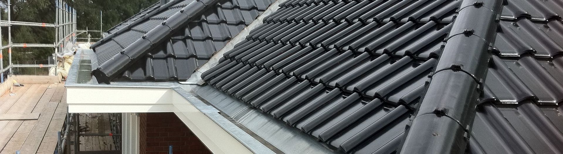Nahaufnahme eines dunkelgrauen Ziegeldachs mit Dachrinne und einem Gerüst im Hintergrund.