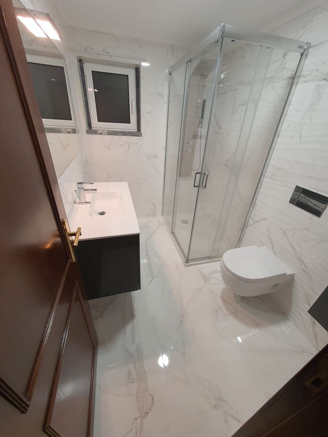 Pequeno banheiro branco com box de vidro, pia com gabinete, vaso sanitário e piso com aparência de mármore.