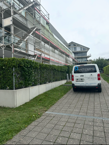 Une camionnette blanche est garée à côté d'un échafaudage et d'une haie, le long d'un chemin pavé près d'un bâtiment.