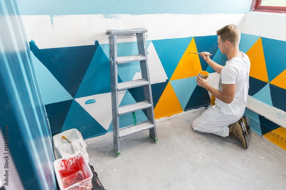 Une personne peint un mur géométrique bleu, jaune et blanc à côté d'une échelle.