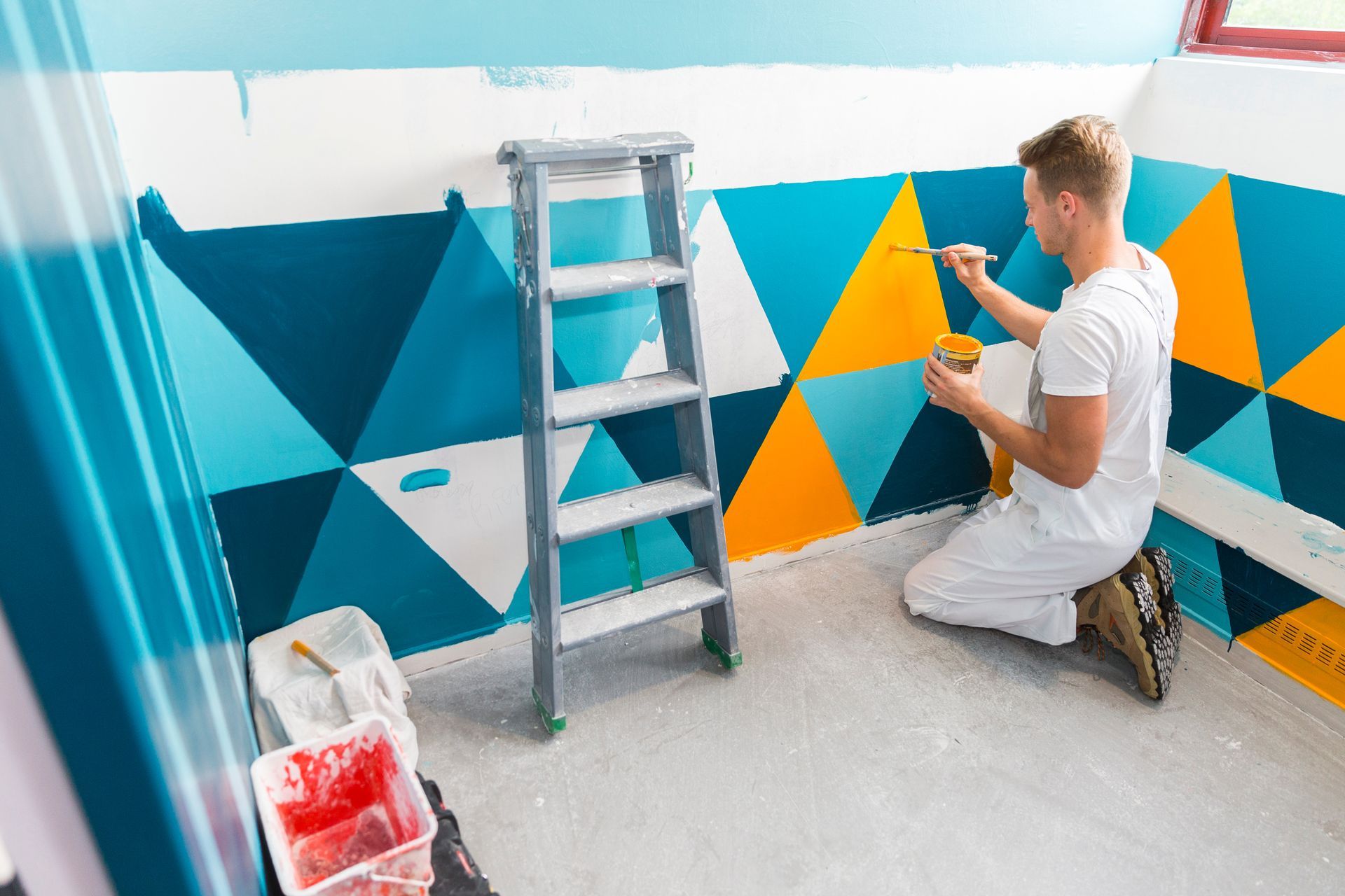 Une personne peint un mur géométrique bleu, jaune et blanc à côté d'une échelle.