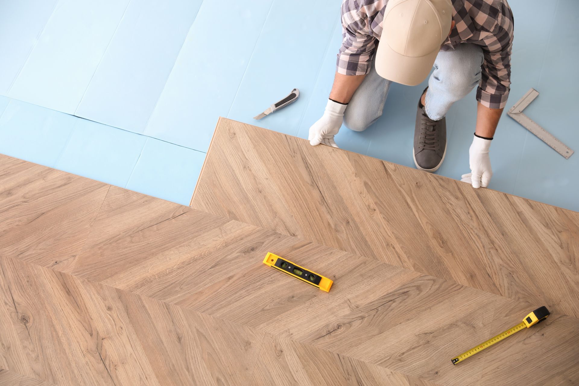 Une personne agenouillée sur un parquet à chevrons, mesurant des planches avec un mètre ruban près d'un mur bleu.