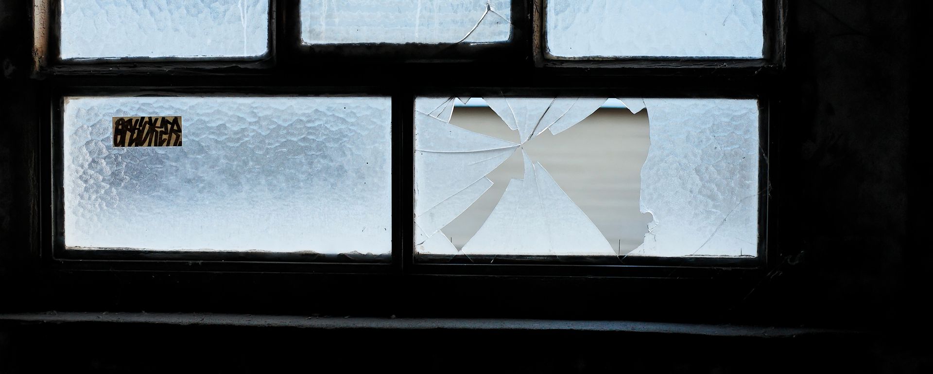 Une fenêtre à croisillons avec une vitre fissurée, laissant apparaître un mur intérieur blanc à travers l'ouverture.