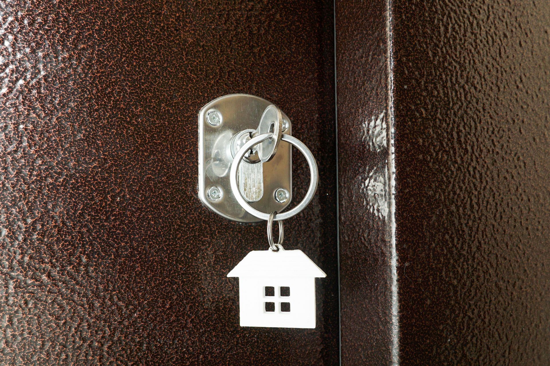 Une clé argentée munie d'un porte-clés en forme de maison est insérée dans une serrure d'une porte en métal texturé brun foncé.