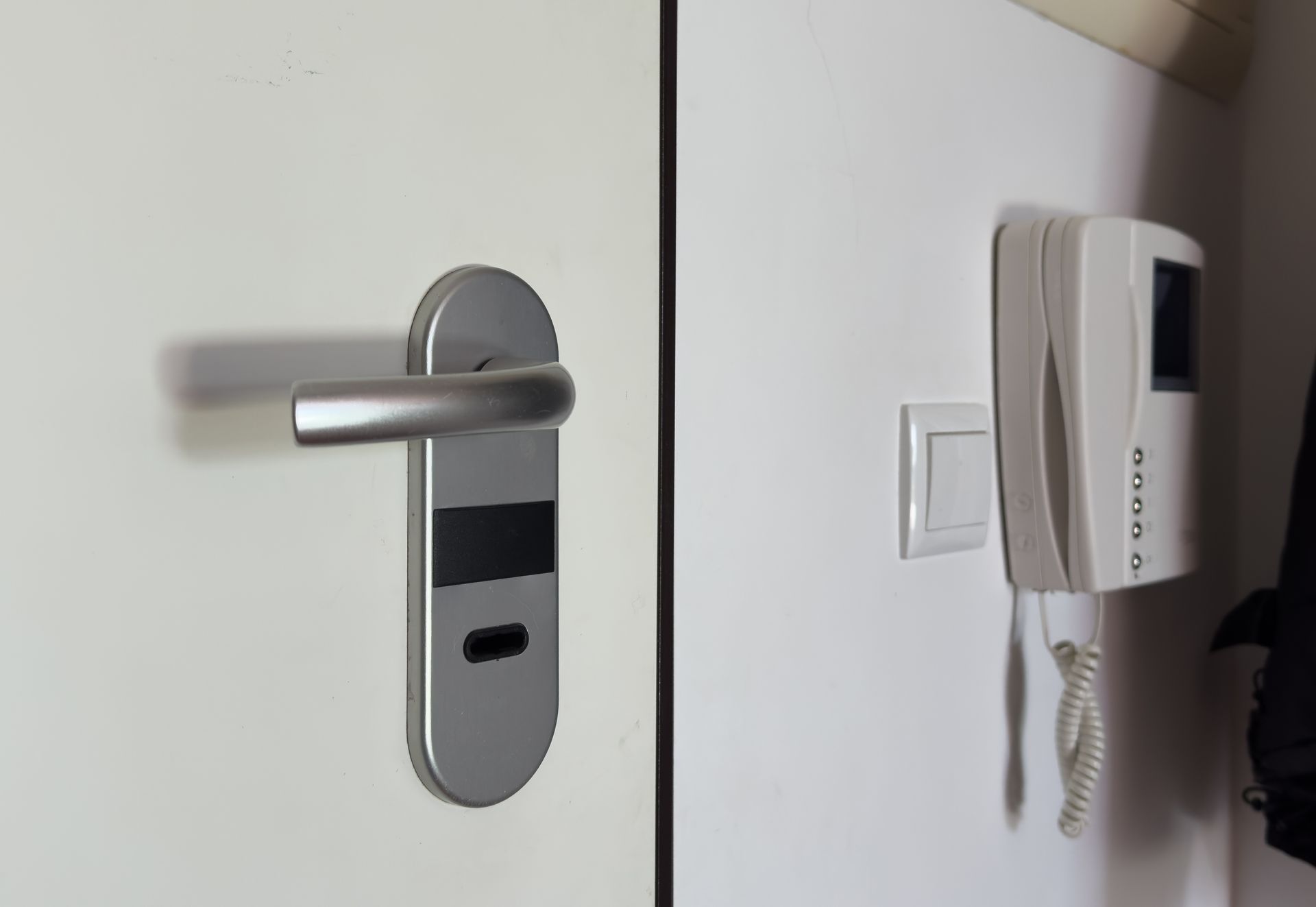 Une poignée de porte argentée avec une serrure à lecteur de carte à côté d'un interphone blanc fixé au mur, sur une porte et un mur blancs.