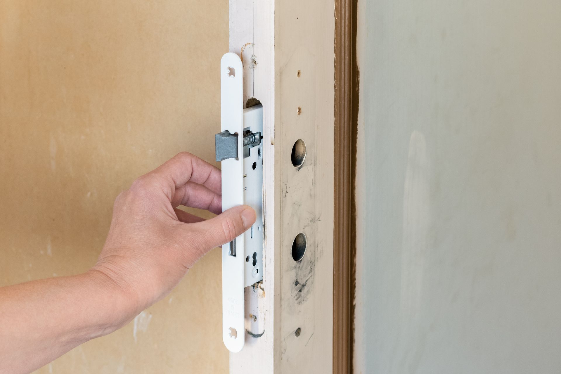 Une main maintient un mécanisme de verrouillage de porte en métal contre le bord d'un cadre de porte en bois, en l'alignant avec les trous de fixation.