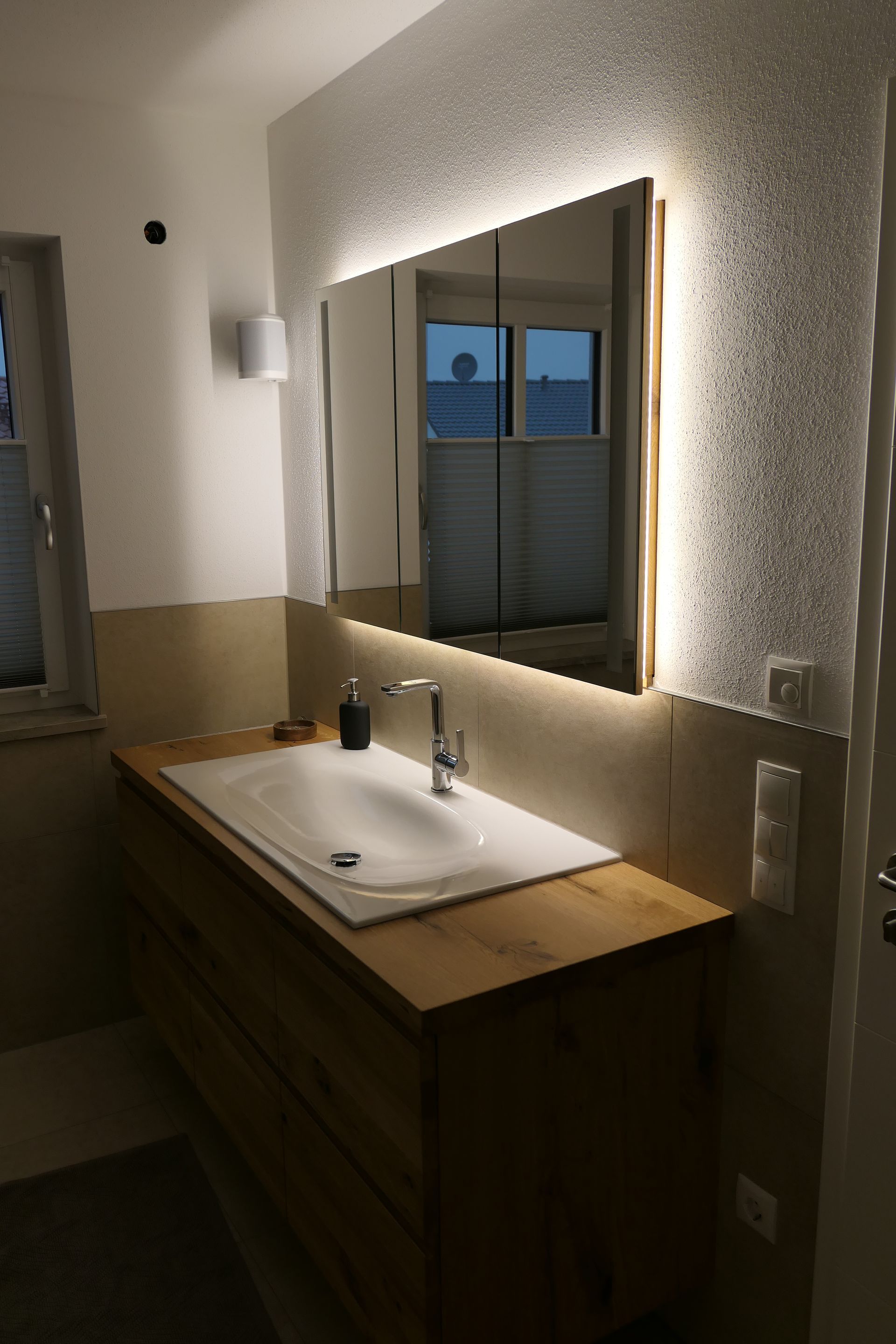 Waschtischunterschrank mit Waschbecken, Spiegel und gedämpfter Wandleuchte neben einem Fenster und einer Holzarbeitsplatte