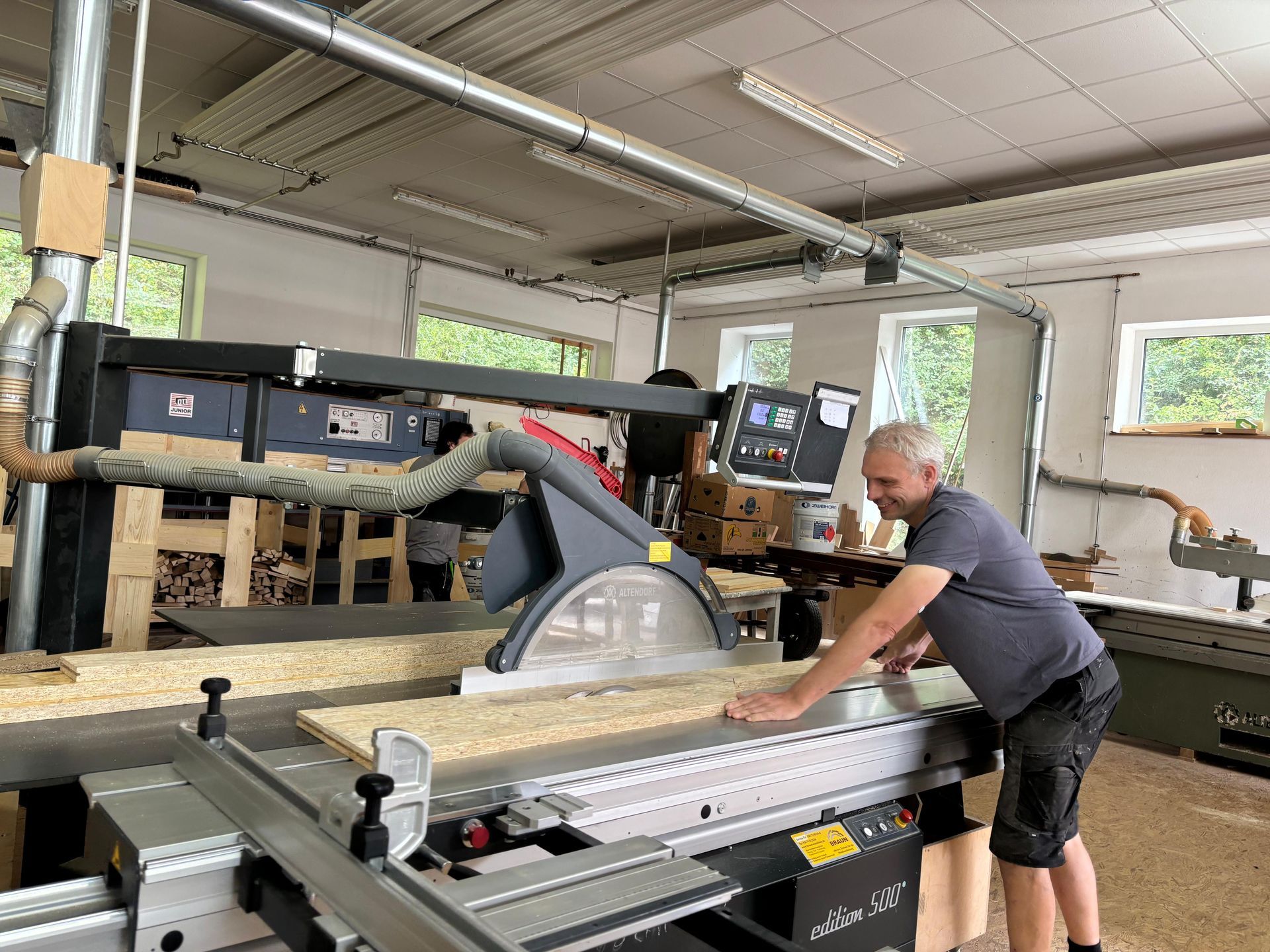 Ein Mann sägt in einer Werkstatt mit Holzbearbeitungsmaschinen und Fenstern ein langes Brett auf einer großen Säge.