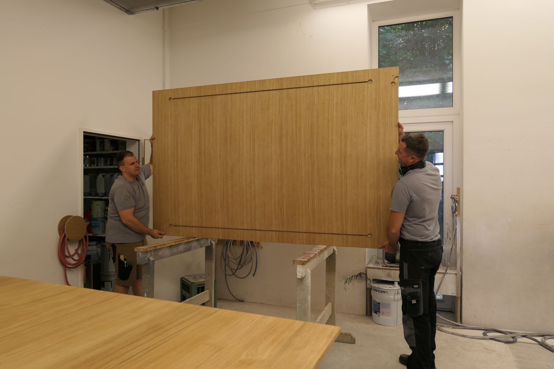 Zwei Arbeiter bewegen eine große Holzplatte in einem Raum, der wie eine Werkstatt aussieht