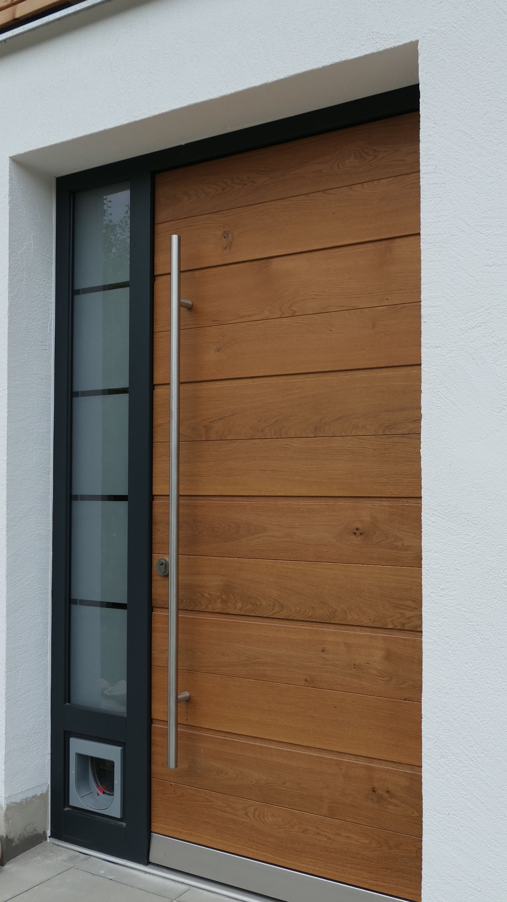 Moderne Haustür mit Holzverkleidung, schwarzem Rahmen und langem silbernem Griff an einer weißen Außenwand
