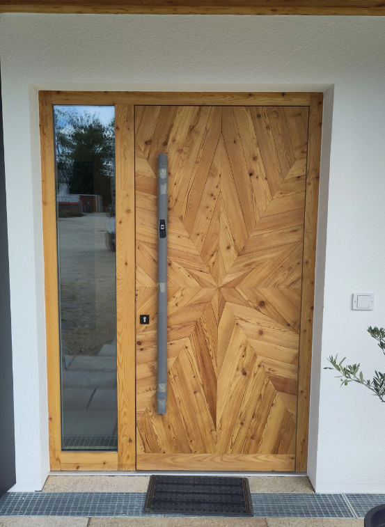 Holzeingangstür mit geometrischem Design, seitlichem Glaselement und schwarzer Fußmatte am Eingang eines modernen Hauses
