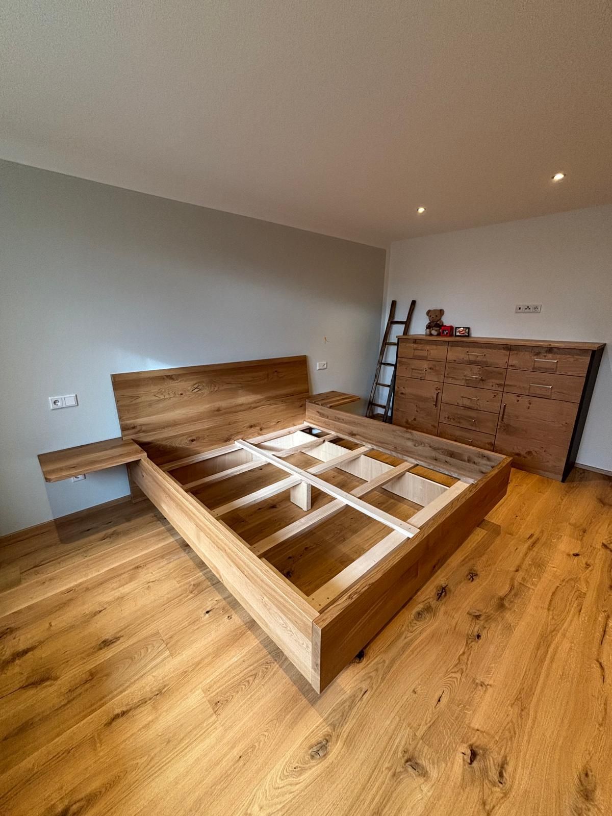 Unfertiger Bettrahmen aus Holz in einem spärlich eingerichteten Zimmer mit Holzboden und einer Kommode in der Ecke.