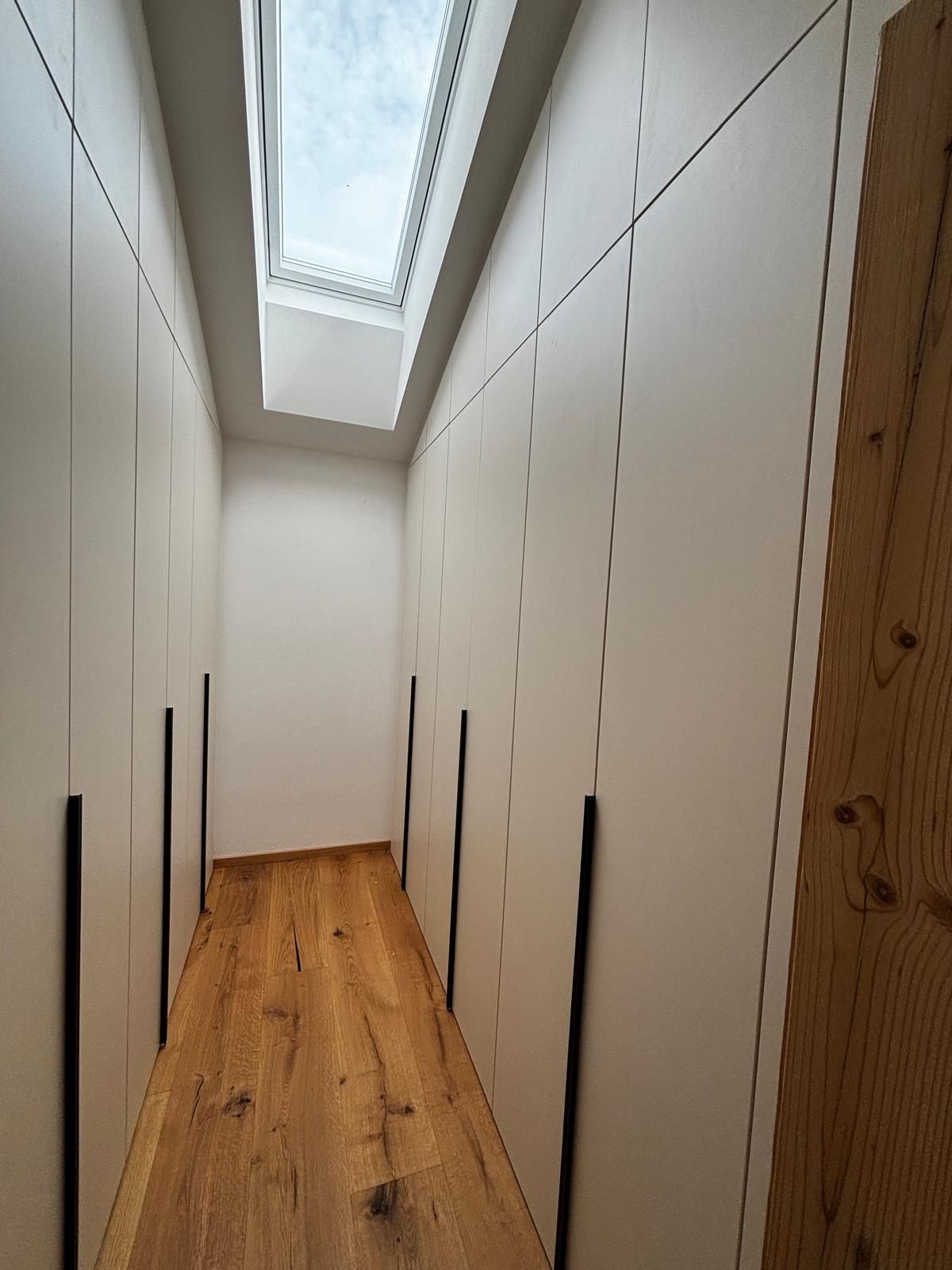 Schmaler Flur mit weißen Wänden, Holzboden und einem Oberlicht an der Decke