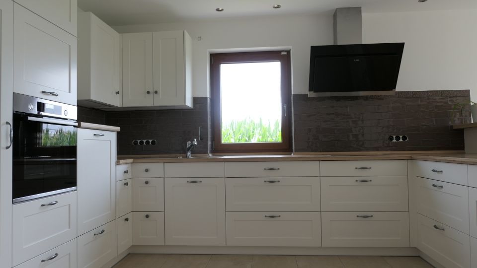 Moderne weiße Küche mit Schränken, schwarzer Rückwand, Fenster und Dunstabzugshaube