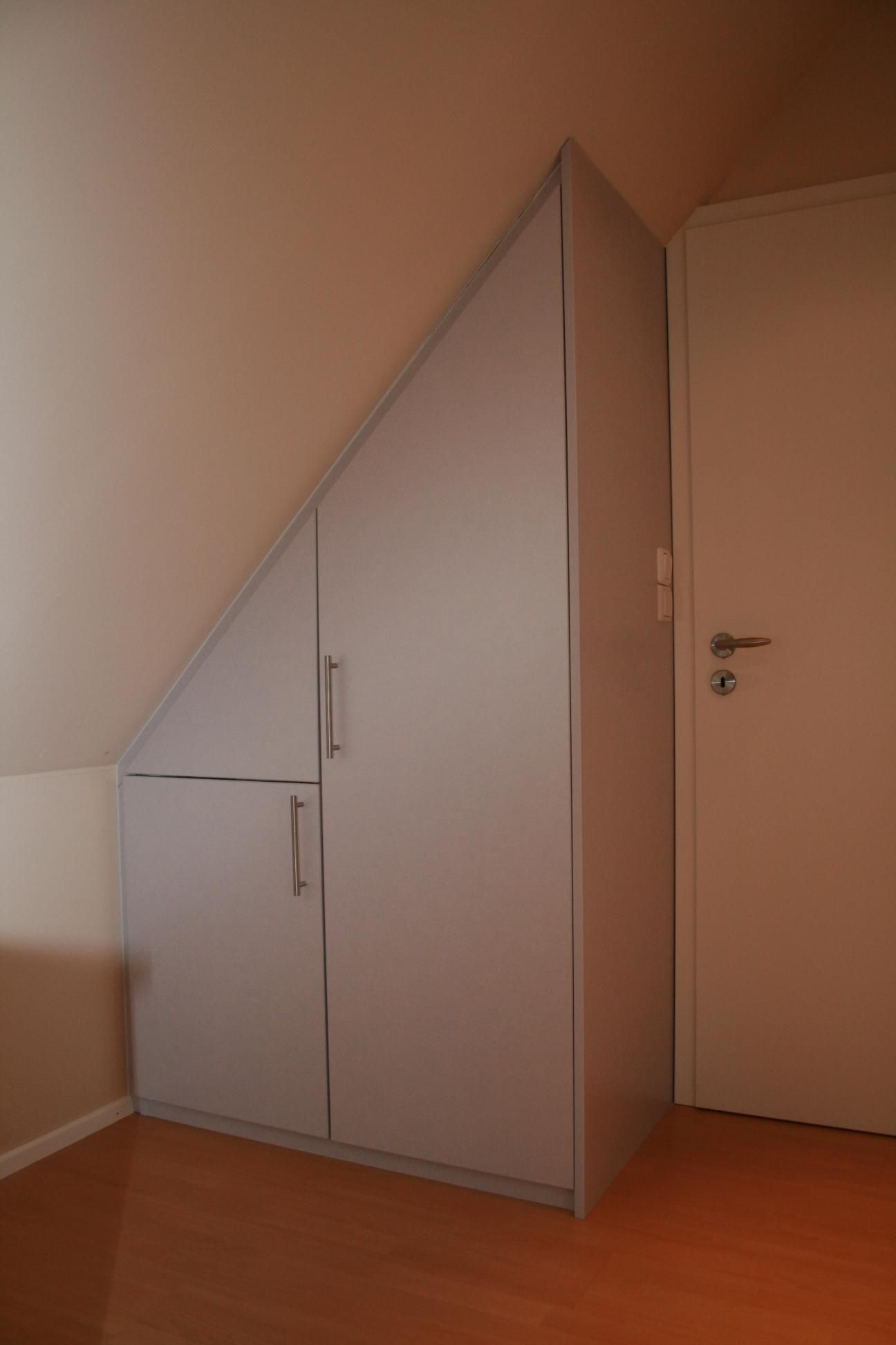 Kleiner dreieckiger Einbauschrank mit grauen Türen in einem Dachzimmer neben einer weißen Tür.