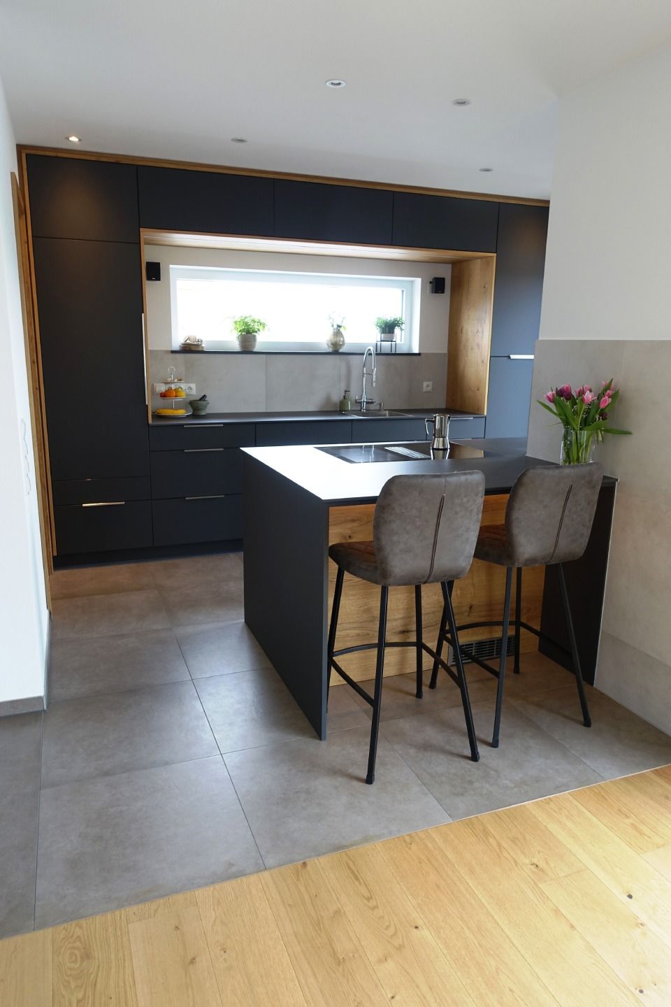 Moderne Küche mit dunklen Schränken, weißer Kücheninsel, zwei Hockern und einem Fenster über der Spüle