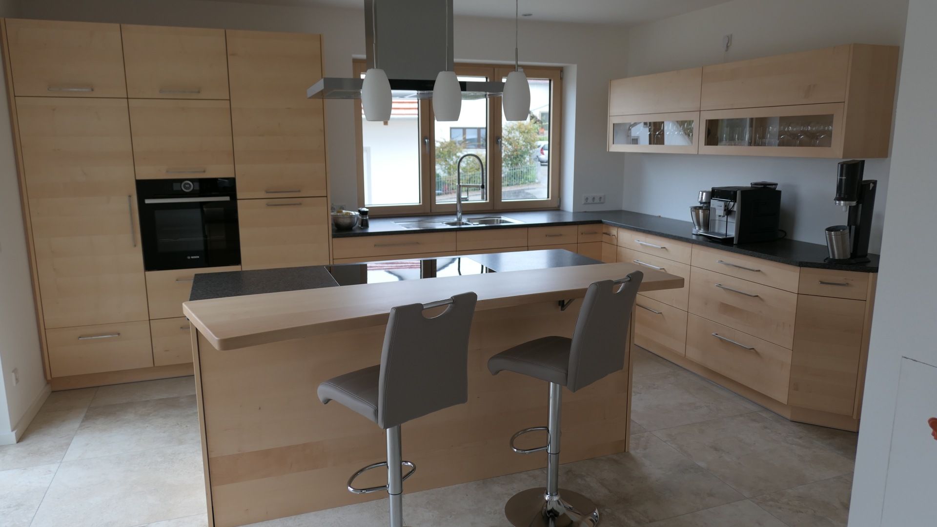 Moderne Küche aus hellem Holz mit Kochinsel, zwei Hockern, Einbaubackofen und Fenster über der Spüle