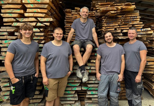 Fünf Personen in grauen Hemden posieren vor gestapelten Holzbrettern, eine davon sitzt auf dem Stapel.