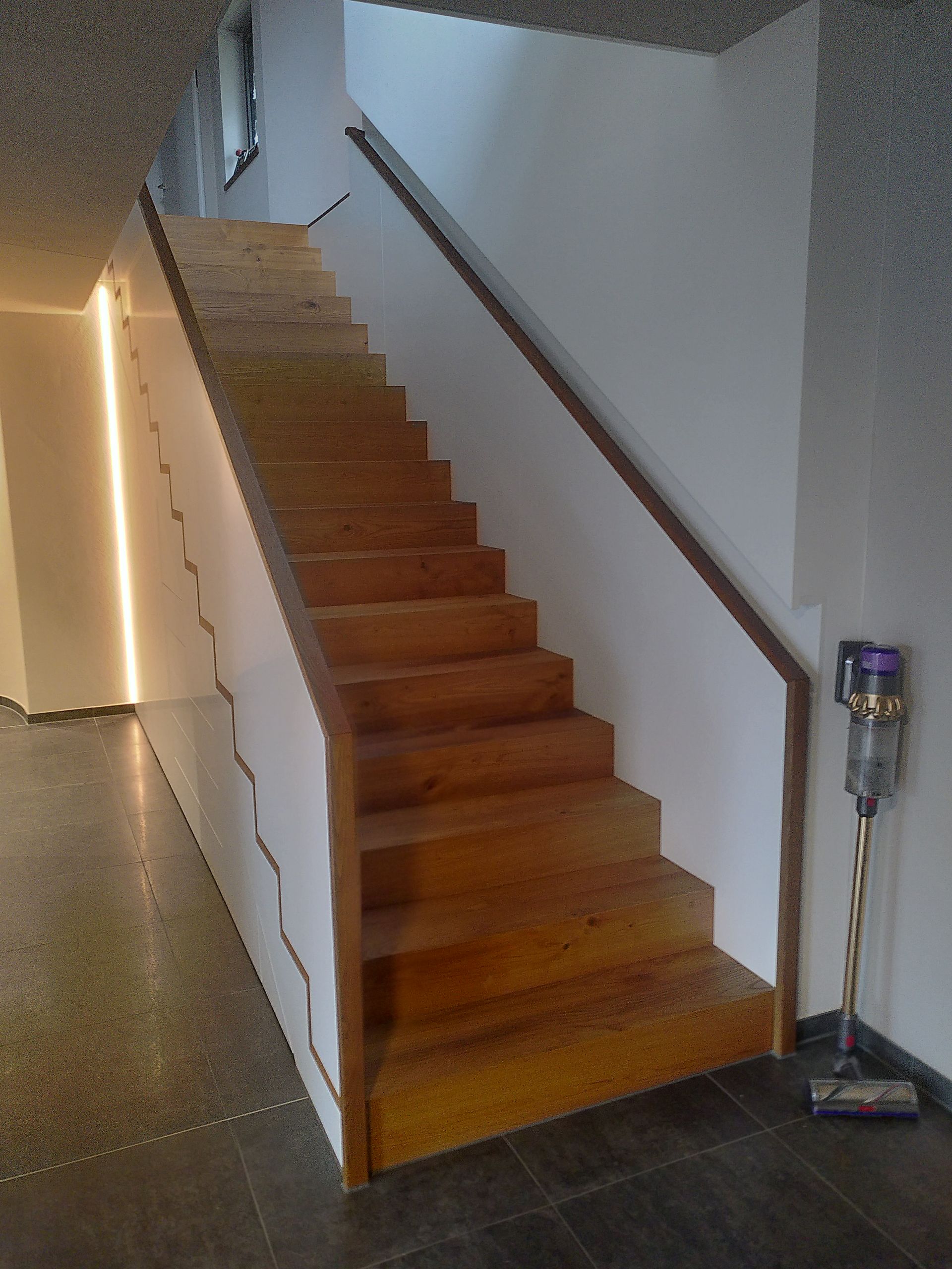 Steile Holztreppe mit weißer Seitenwand und Handlauf in einem schmalen Innenflur