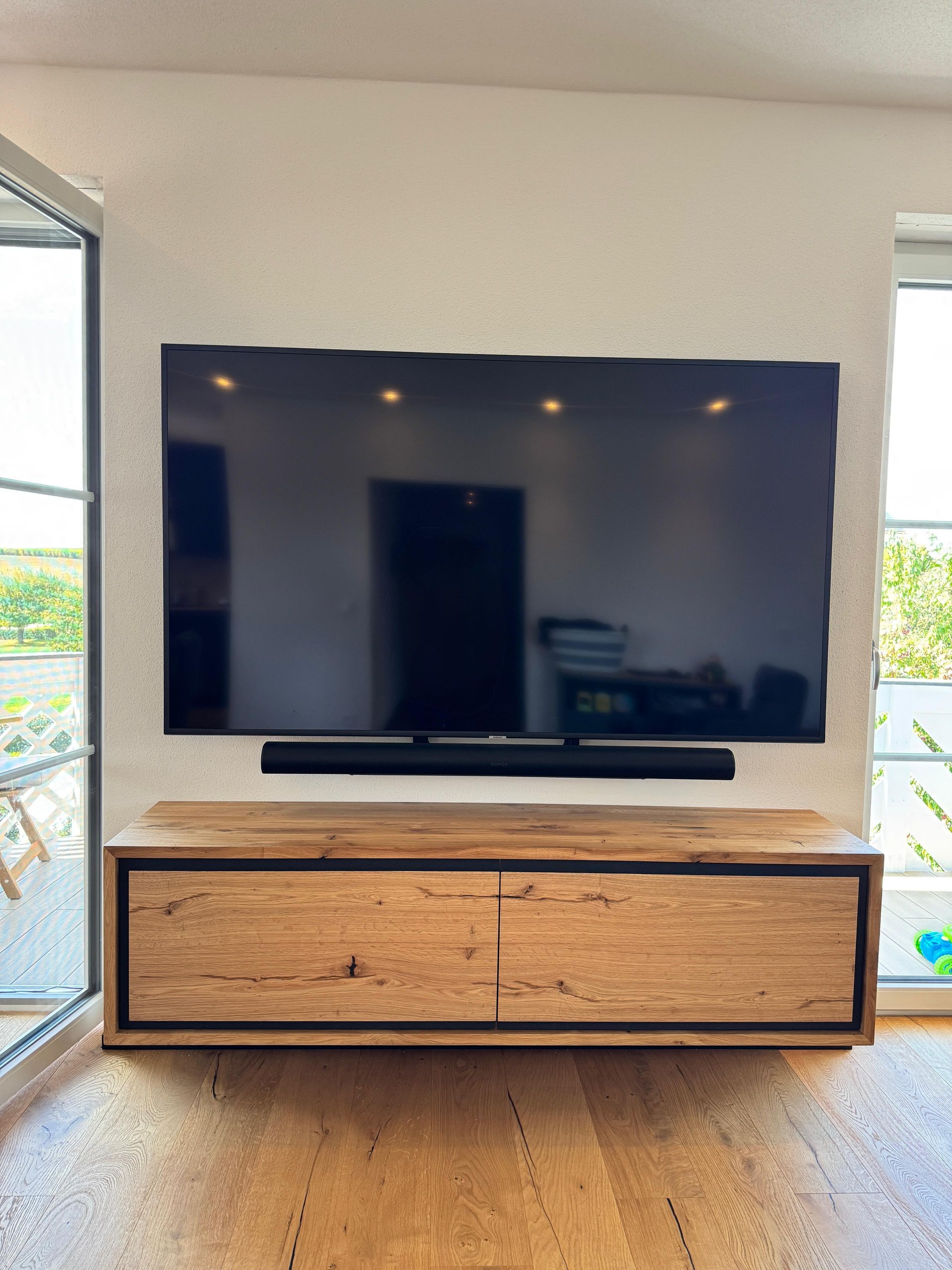 An der Wand montierter Fernseher über einem TV-Lowboard aus Holz in einem hellen Raum mit Fenstern auf beiden Seiten