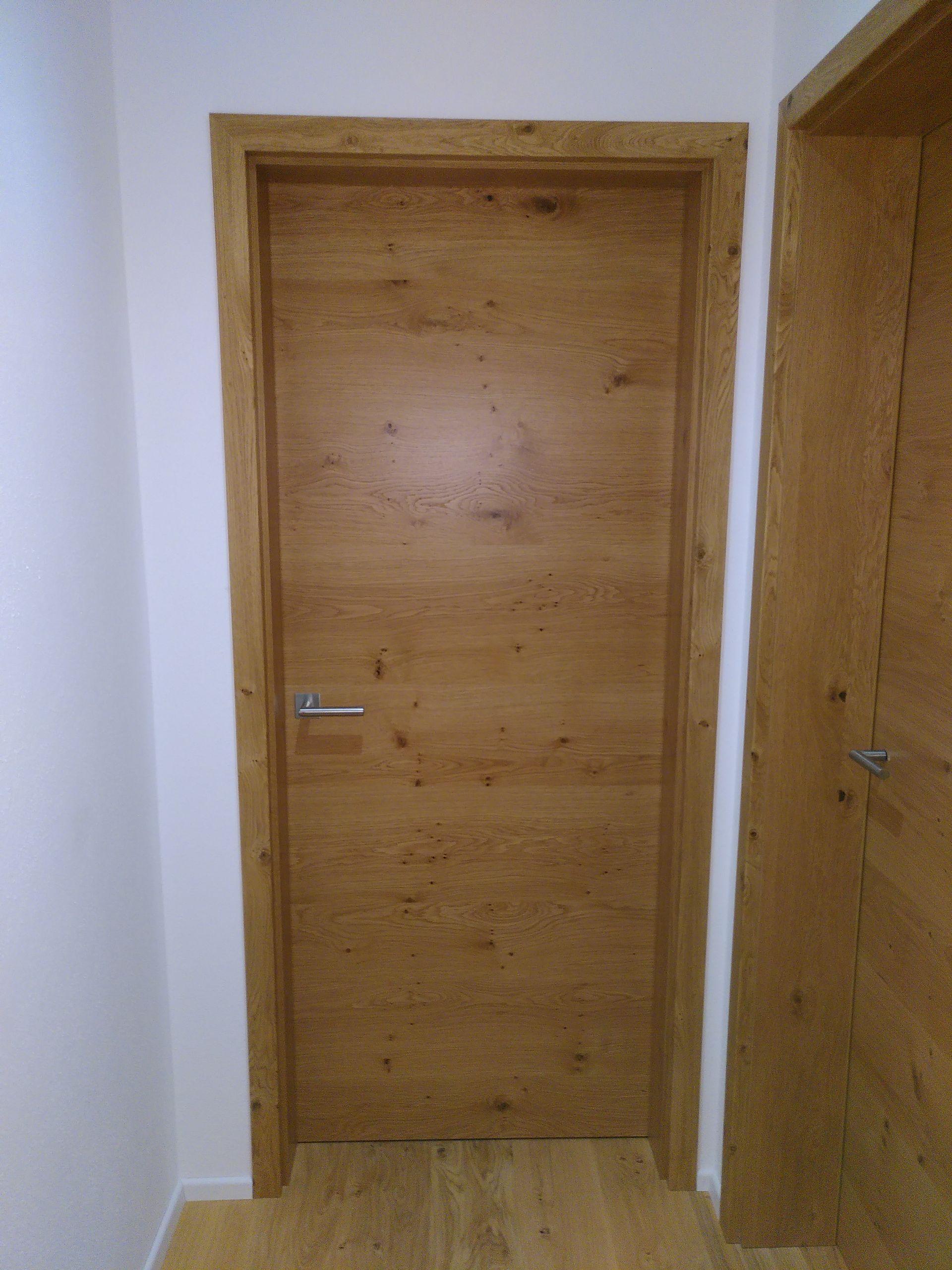 Holz-Innentür in einem weißen Flur, rechts ist eine weitere Tür teilweise zu sehen.