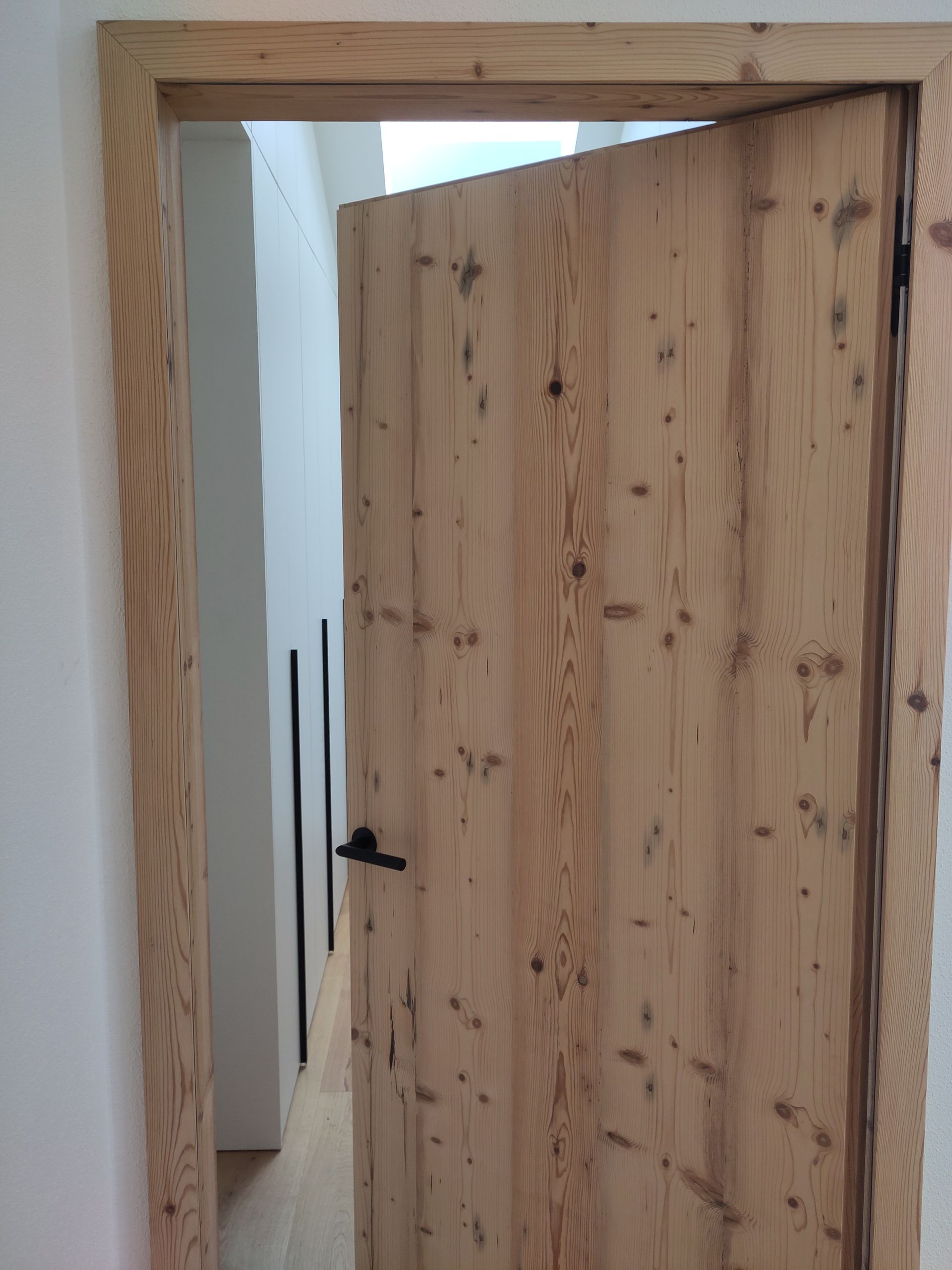 Teilweise geöffnete Holztür in einem schmalen Flur mit weißen Wänden