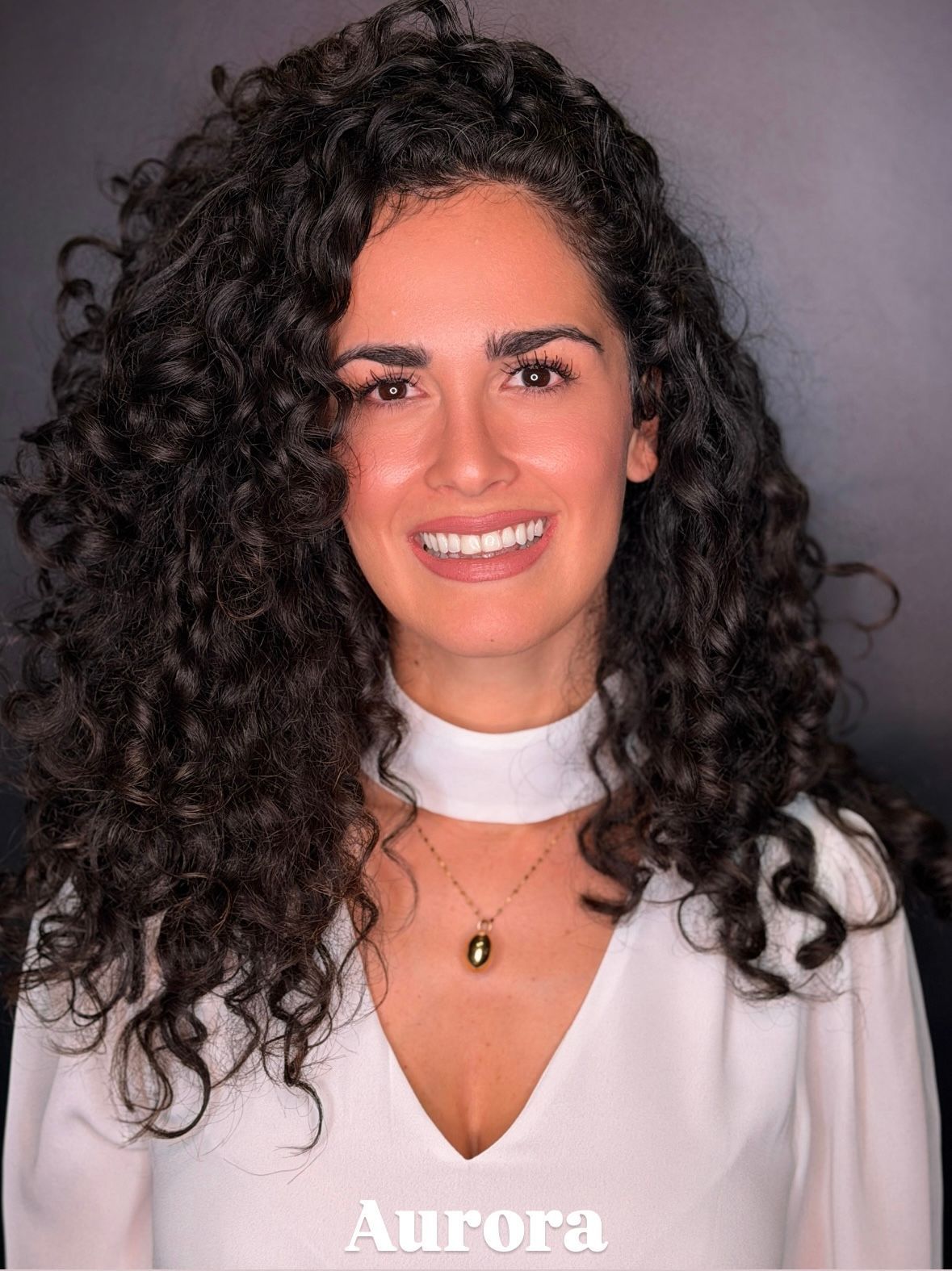Donna sorridente con lunghi capelli scuri e ricci, che indossa una maglietta bianca e una collana con pendente d'oro, su sfondo grigio.