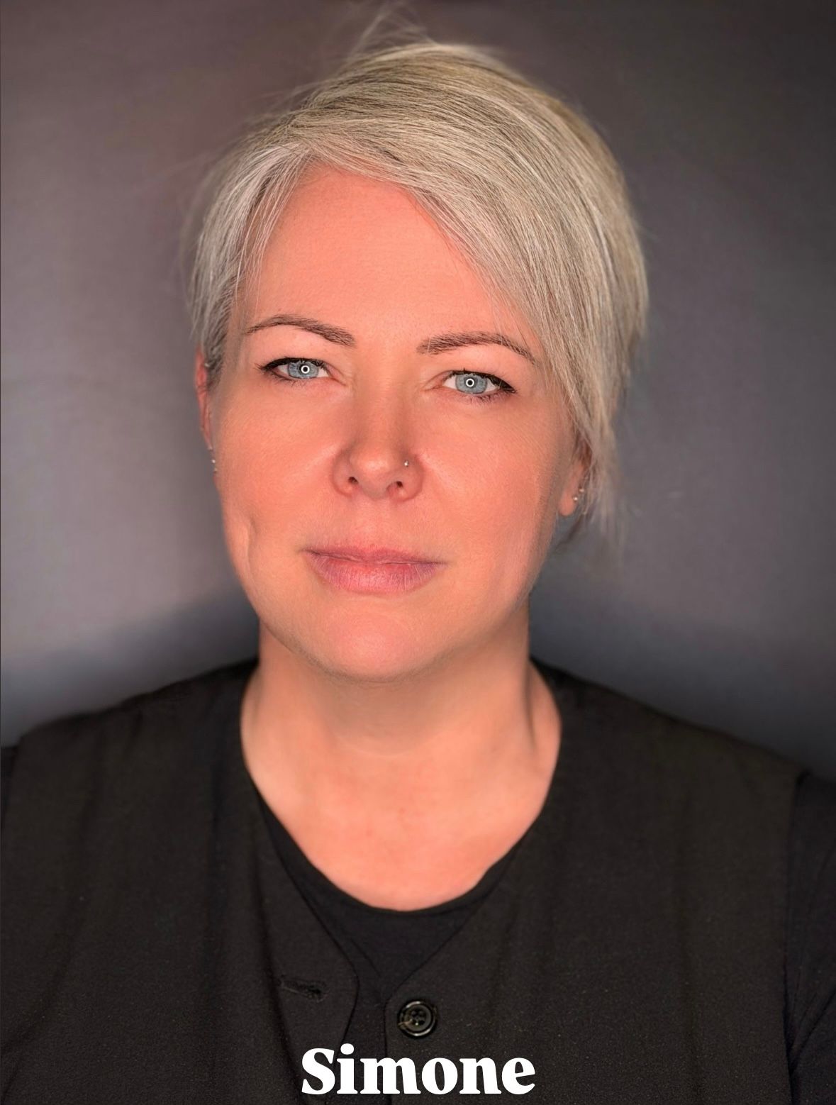 Primo piano di una persona con capelli corti grigi e occhi azzurri, che indossa una camicia nera con colletto su sfondo grigio.