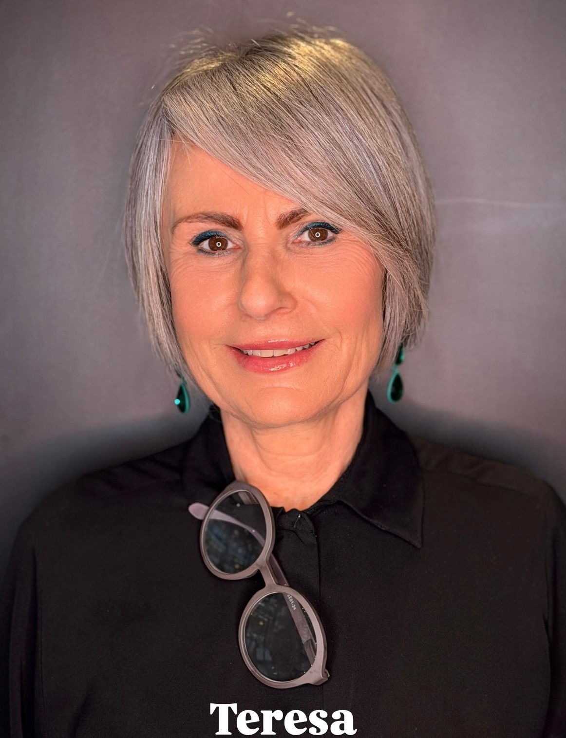 Ritratto di una persona con capelli corti e grigi, che indossa una camicia nera, orecchini verdi e occhiali appesi al colletto.