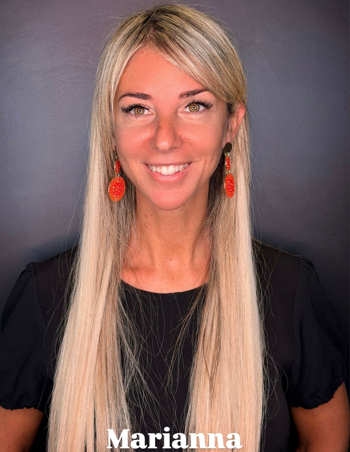 Una persona sorridente con lunghi capelli biondi e orecchini pendenti rossi, centrata su uno sfondo grigio scuro.