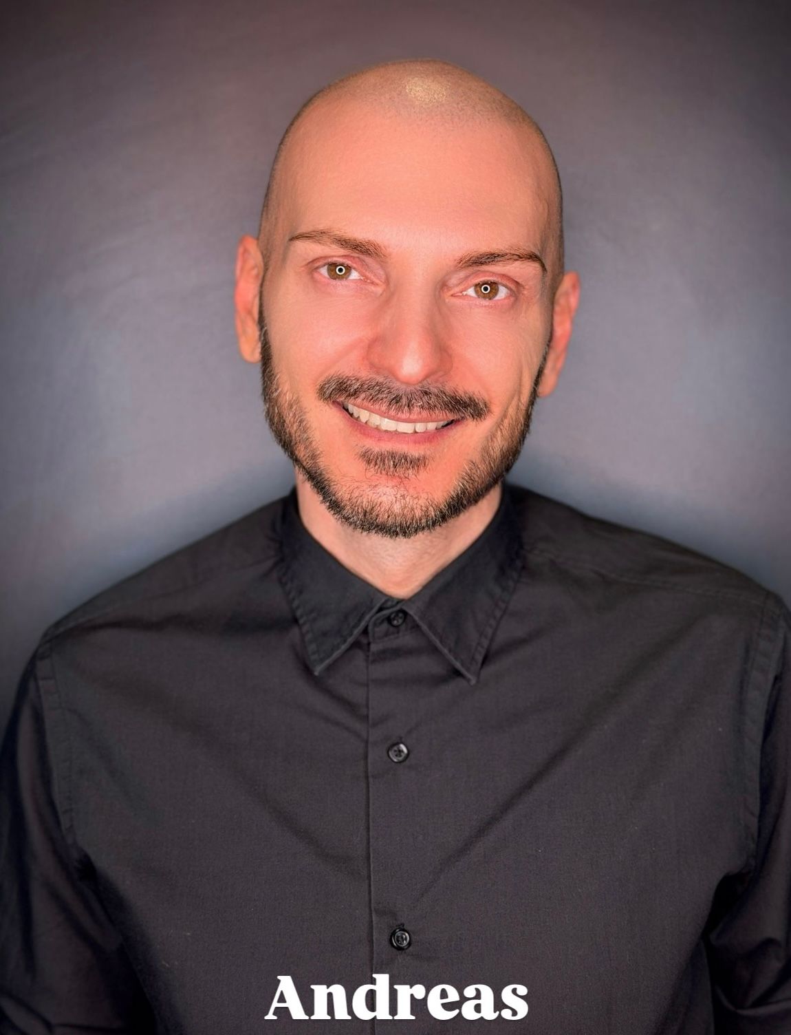 Una persona sorridente con la testa rasata e la barba, che indossa una camicia nera con colletto, su uno sfondo grigio.