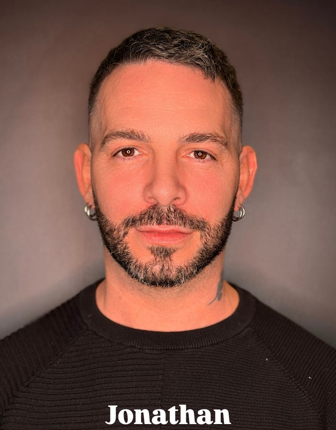 Una persona con capelli corti e scuri, barba e orecchini a cerchio, in posa contro uno sfondo scuro con la scritta