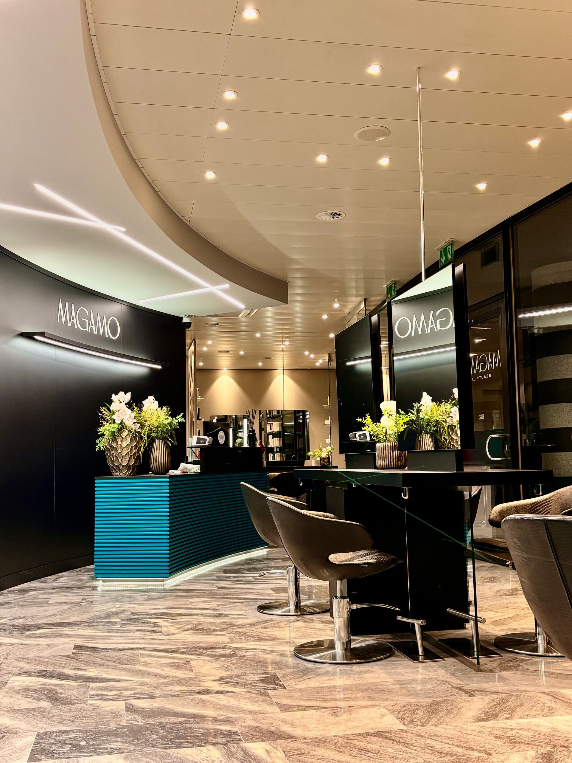 Un moderno salone da parrucchiere caratterizzato da un bancone reception con texture color verde acqua, postazioni di lavoro nere con specchi e pavimento in marmo.