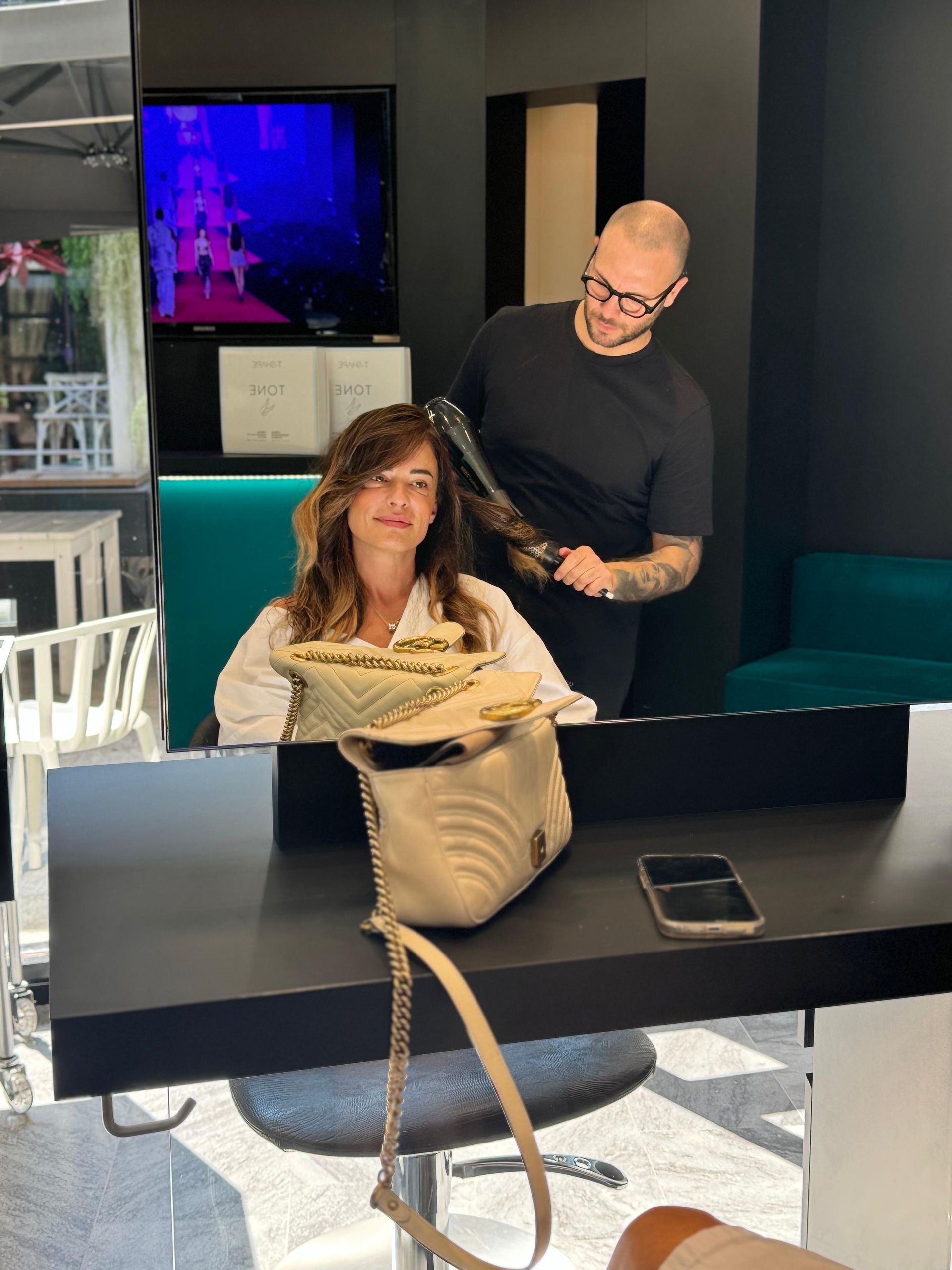 In un salone, una parrucchiera usa un asciugacapelli sui capelli di una cliente, mentre su un bancone nero è appoggiata una borsetta color crema.