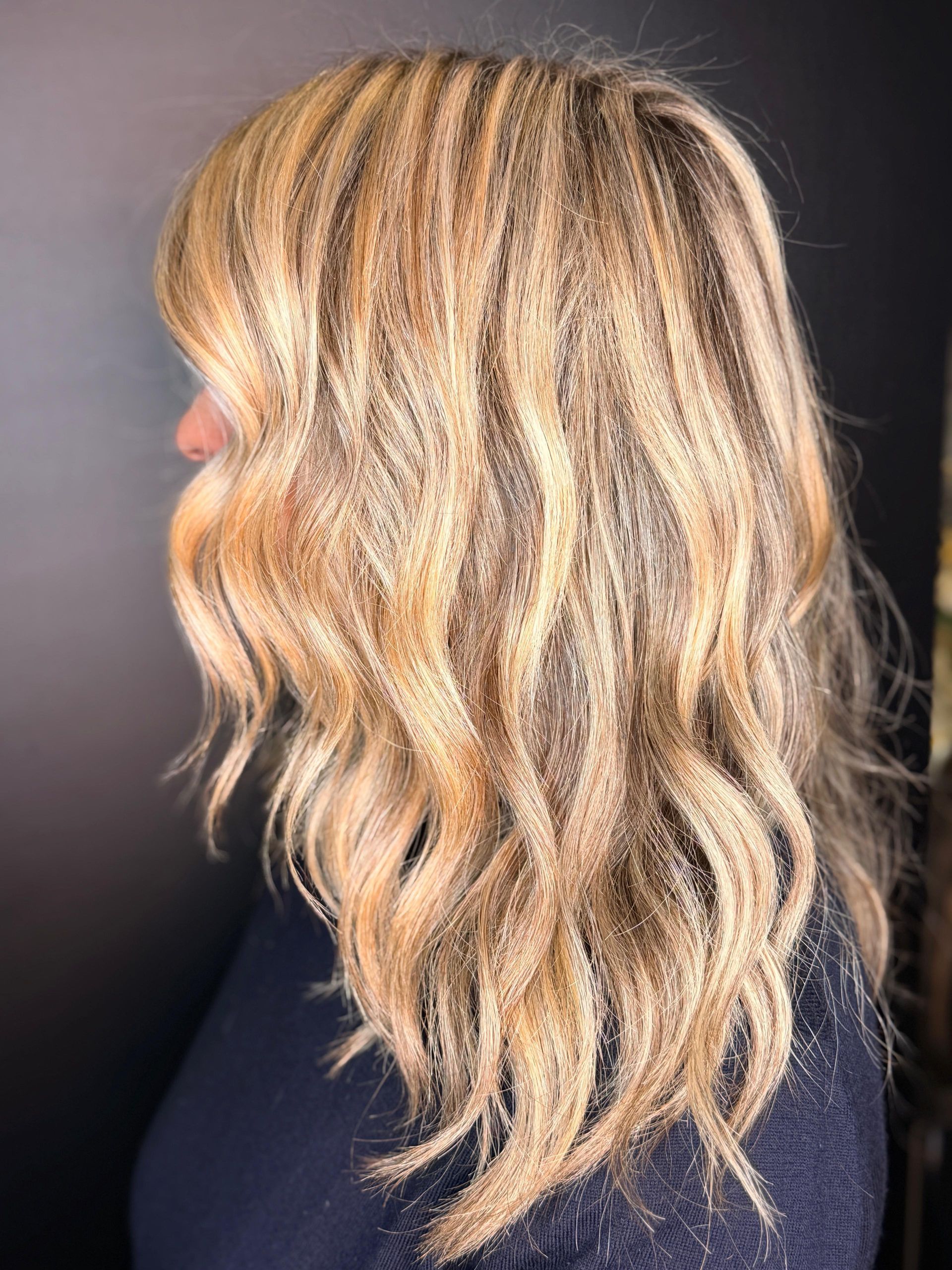 Una vista di profilo di capelli biondi, mossi e con mèches, su uno sfondo scuro.