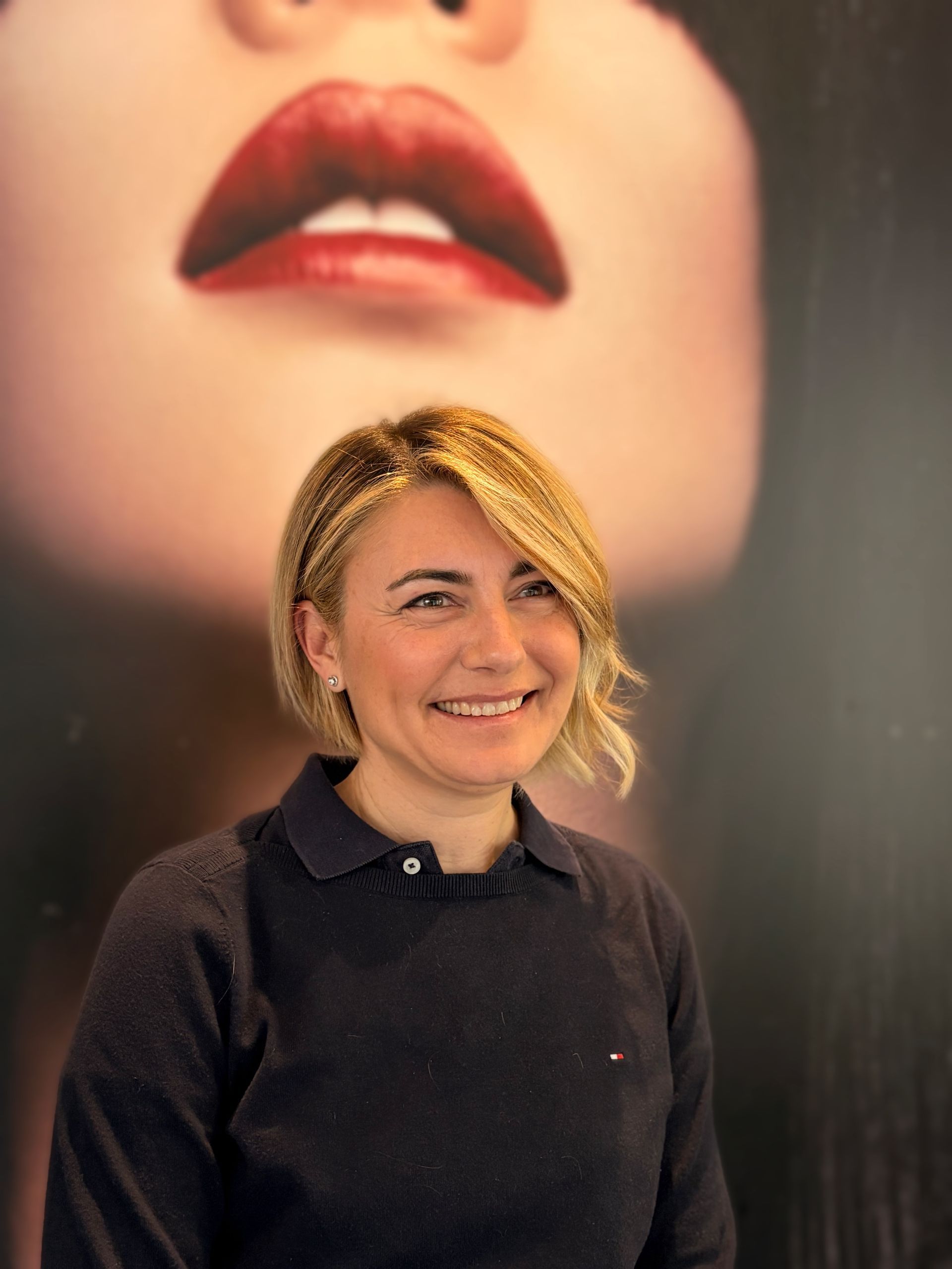 Una persona sorridente con capelli biondi di media lunghezza, che indossa un maglione blu scuro con colletto, posa davanti a un murale raffigurante un volto. MAGAMO Lugano