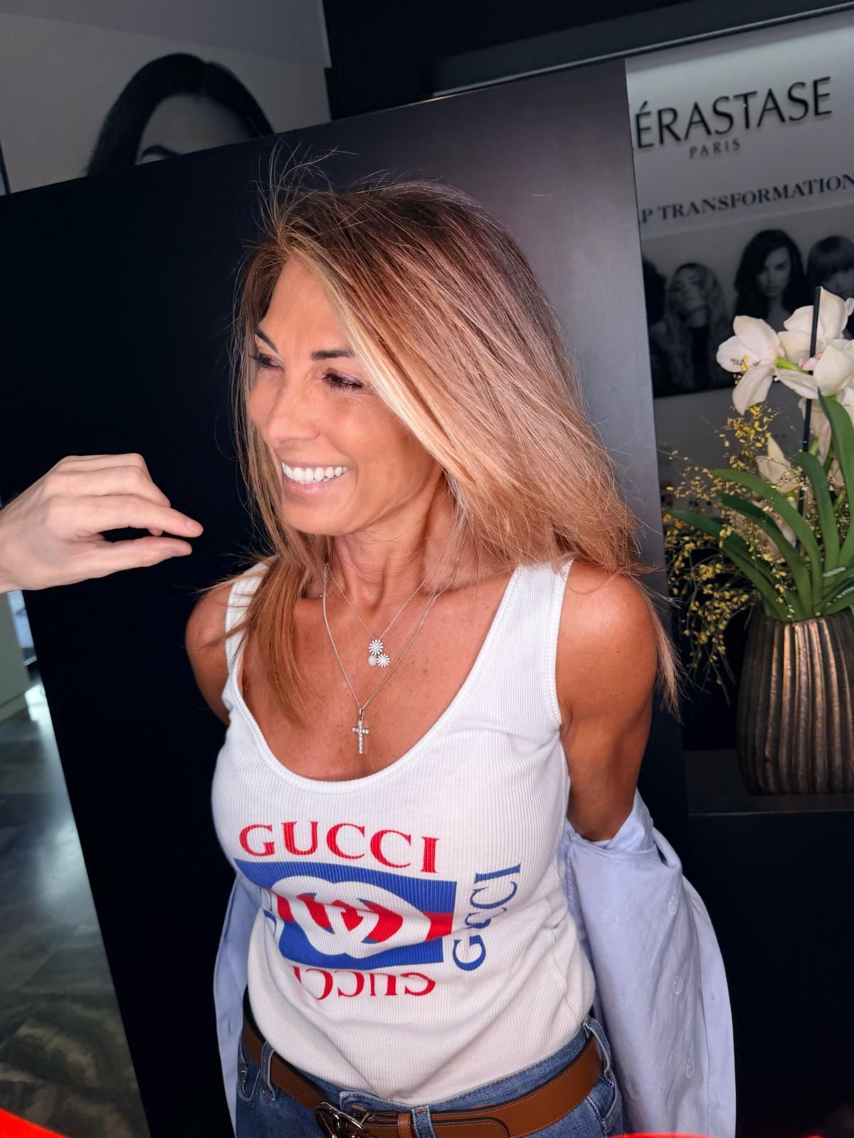 Una persona sorridente con una canotta Gucci e jeans blu è in piedi in un salone e si porta una mano ai capelli.