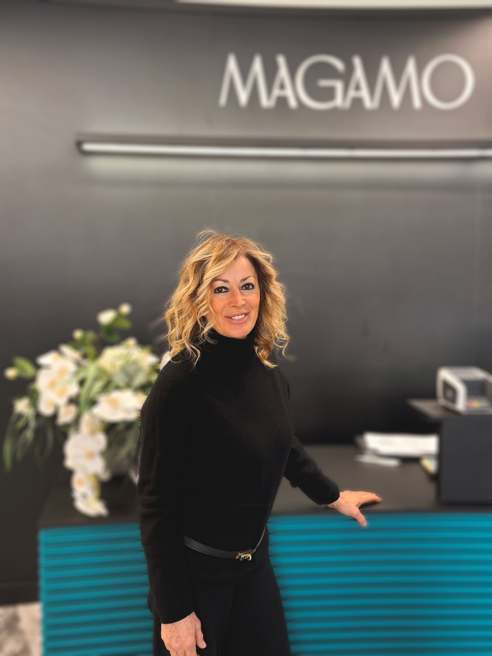 Una donna sorridente con i capelli biondi e ricci è in piedi dietro un bancone turchese, di fronte a una parete con il logo MAGAMO. MAGAMO Lugano