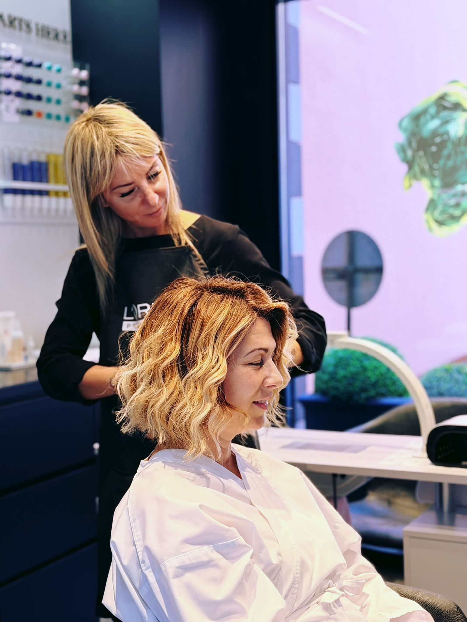 In un salone di parrucchieri luminoso e moderno, una stilista acconcia i capelli mossi e biondi con mèches di una cliente. MAGAMO Lugano, Titolare 