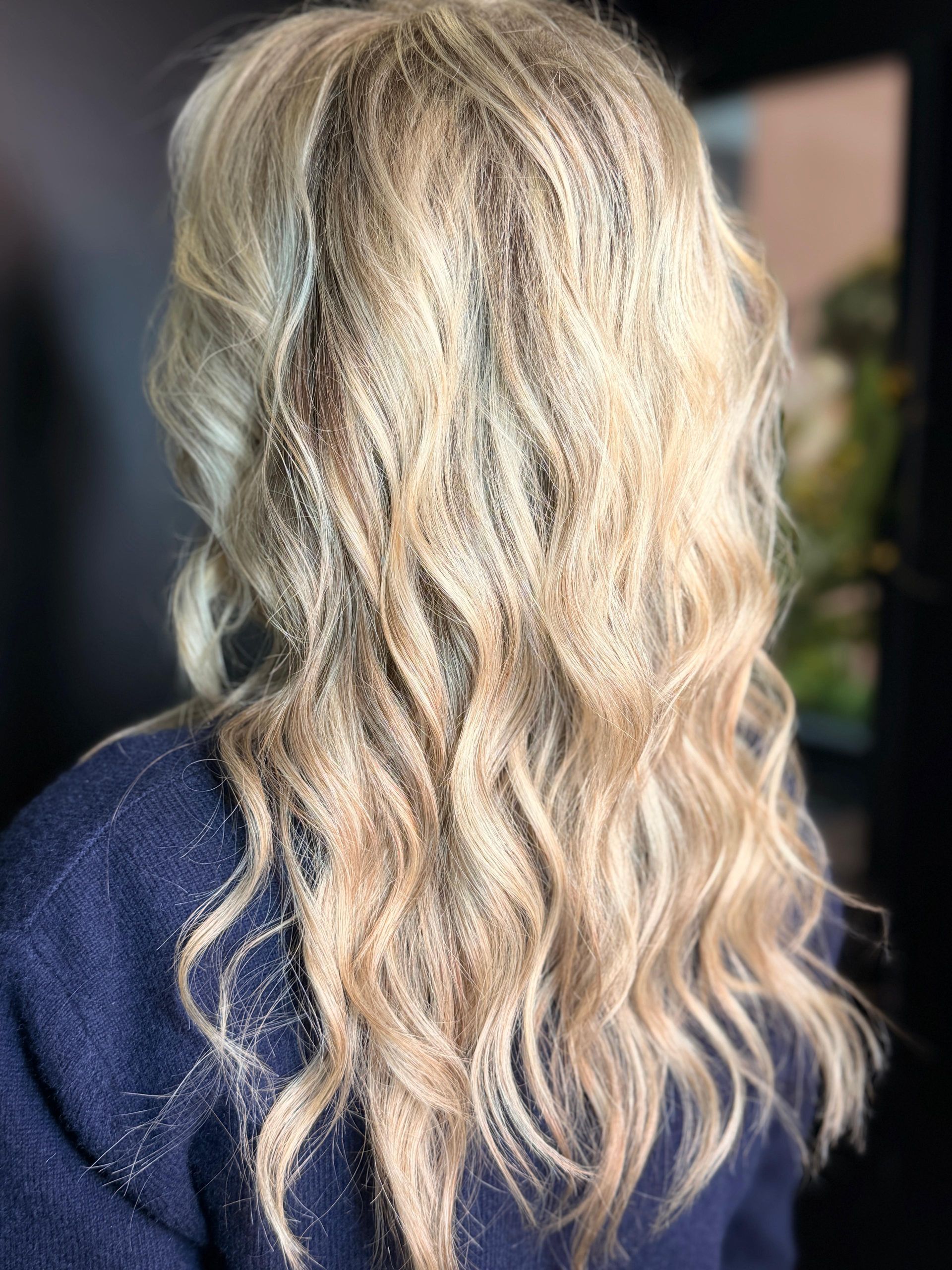 Vista posteriore di lunghi capelli biondi e ondulati, acconciati con morbidi riccioli, su uno sfondo scuro e sfocato.